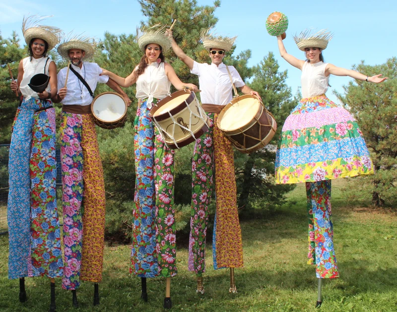 Five TallBeat drummers in white tops and floral fabric pants displaying alfaia drums and cowbell at park festival